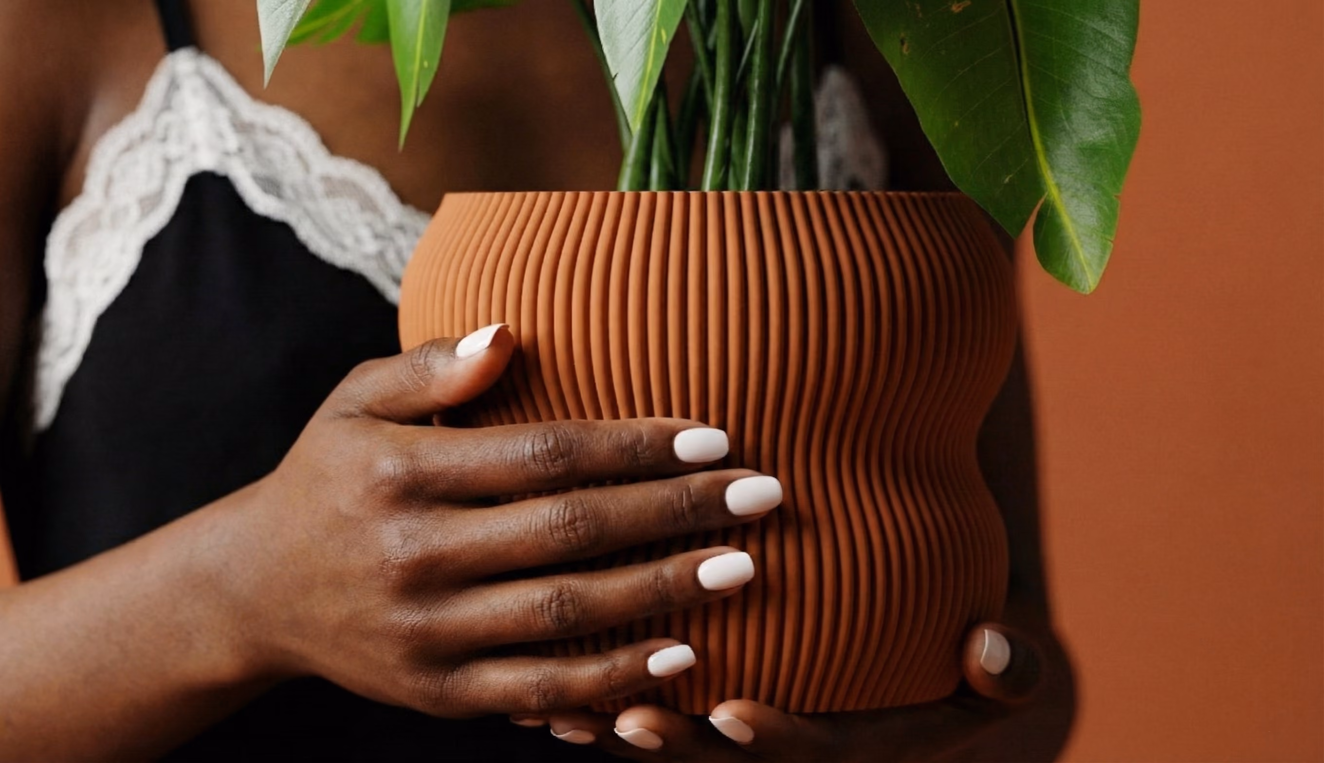 Hands with white nails holding a ribbed brown pot with green leaves against a brown background.